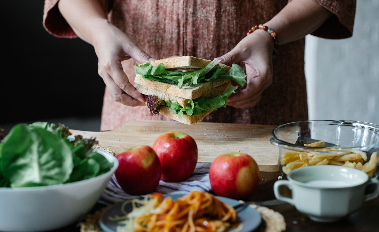 about-us Unrecognizable female standing at table with apples and cutting board with sandwich with lettuce in hands in kitchen on breakfast time